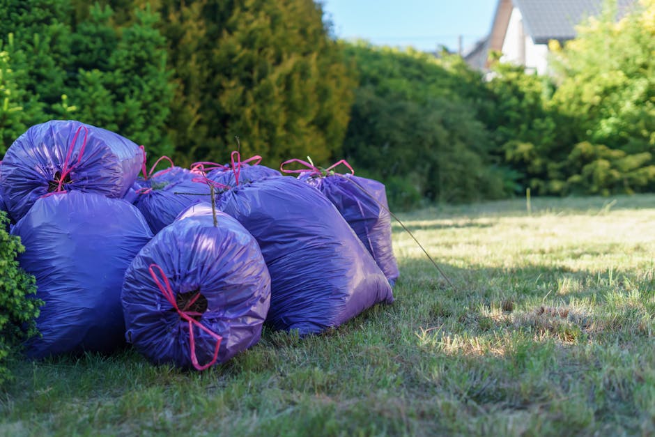 Several purple garden waste sacks tied with pink cords are stacked along the edge of a well-maintained grassy lawn, adjacent to a shrubbery with lush green foliage. The grass exhibits a mix of trimmed