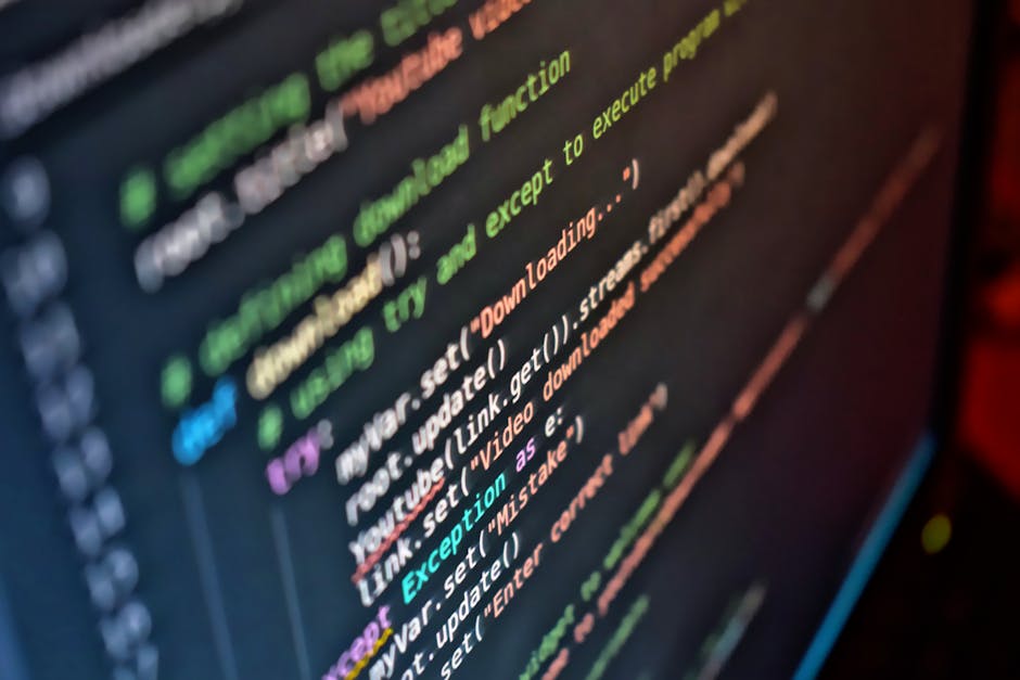 A close-up photograph of a computer screen displaying lines of colorful programming code with syntax highlighting in shades of green, blue, purple, orange, and white on a dark background. The code app